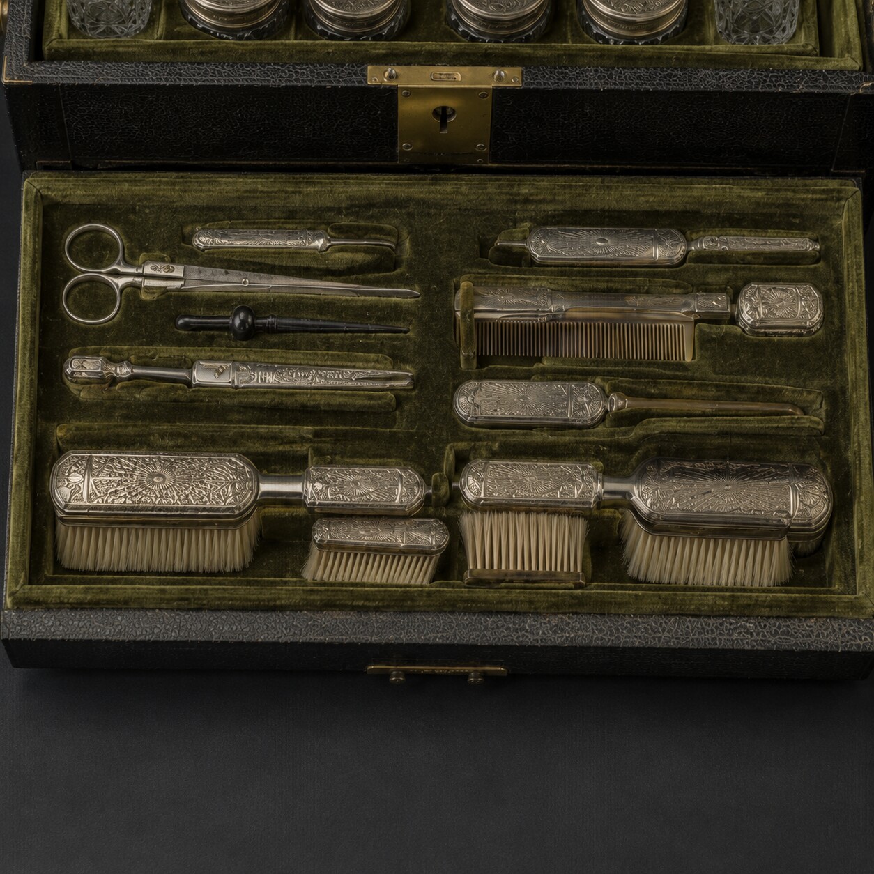 Overhead editorial reconstruction of silver-mounted grooming tools arranged in fitted olive velvet recesses within a black leather tray.
