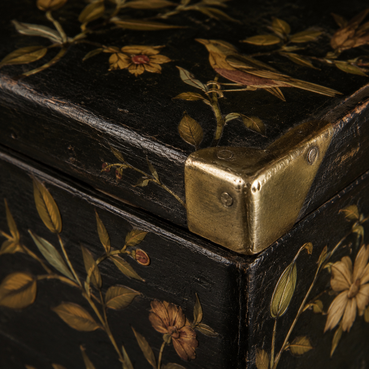 Close view of floral and bird inlay on black lacquer beside an aged brass corner fitting.